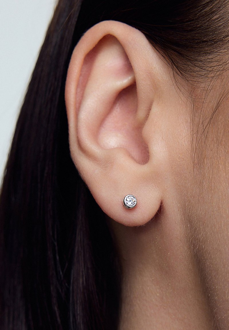Srebrna kolczyk w kształcie okręgu, z przezroczystym kamieniem w centrum. Ujęcie zbliżeniowe ucha.