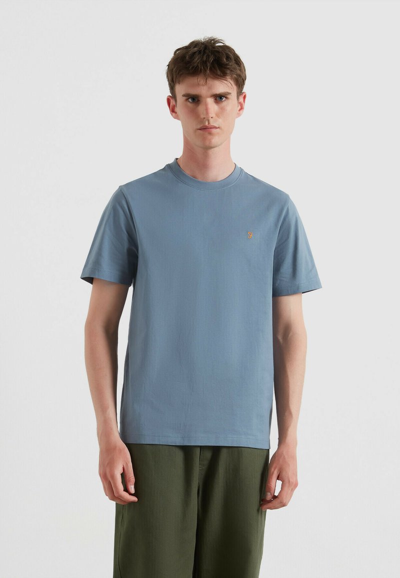 T-shirt di cotone azzurro chiaro con maniche corte e collo rotondo, caratterizzata da un piccolo logo giallo ricamato sul petto. Indossata con pantaloni verdi.