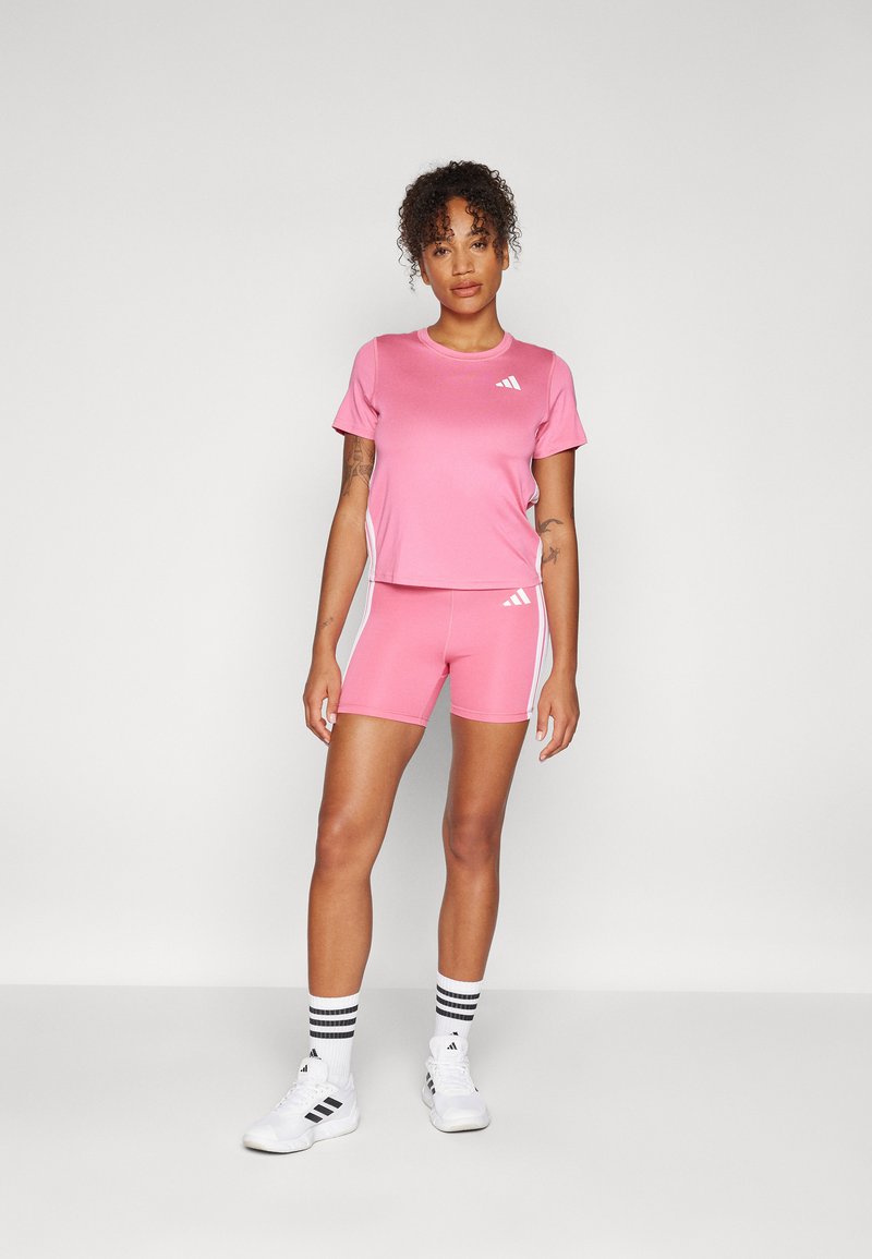 Ensemble de sport rose comprenant un haut à manches courtes et un short ajusté, tous deux ornés d'accents blancs et d'un logo. Le matériau est lisse et extensible.