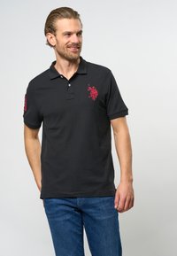 Juodas polo marškinėliai iš medvilnės mišinio, su raudonu siuvinėtu logo ant krūtinės ir raudonu akcentu ant rankovės. Trumpomis rankovėmis, klasikinio apykaklės tipo.