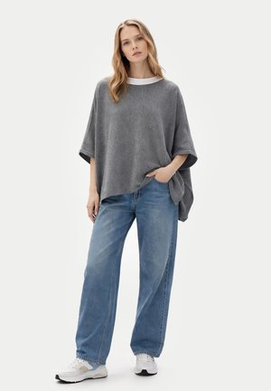 Jeune femme debout, main dans la poche, portant un poncho gris ample, un jean large bleu clair et des baskets blanches sur fond uni.