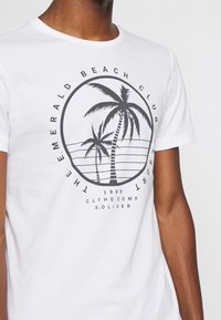 Camiseta de algodón blanca con un gráfico circular negro que presenta palmeras y el texto: "THE EMERALD BEACH CLUB" y "1969". Mangas cortas, cuello redondo.