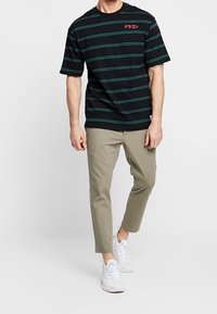 T-shirt nera a maniche corte con strisce verdi, con testo rosso sul petto. Abbinata a pantaloni tapered color khaki chiaro e sneakers bianche.