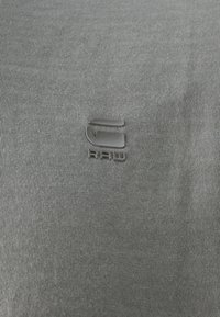 Pilkas audinys su lygiu, tekstūruotu paviršiumi. Pasižymi ryškiu juodu logotipo, su tekstu "RAW" po juo, centruotu ant audinio.