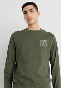 Olīvu zaļš t-krekls ar garām piedurknēm, izgatavots no kokvilnas. Iekļauj apaļu kakla izgriezumu un priekšējo grafiku ar "RB/20" baltā un dzeltenā krāsā. Gluda virsma.