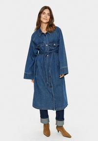 Jeansový dlhý kabát s pásom v páse, zapínaním na gombíky, dvoma prednými vreckami, širokými rukávmi a modrým praním. Styling s vykasanými džínami a hnedými topánkami.