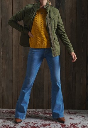 Person iført grøn jakke, sennepsgul kabelstrikket sweater, udslåede blå jeans og brune sko, stående foran en væg af træ.