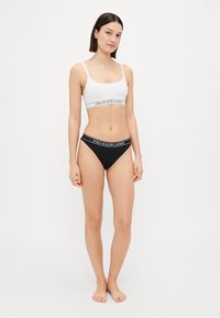 Hvit bralette med et svart elastisk bånd som har "POLO RALPH LAUREN" logo, kombinert med svarte bikinibukser med lignende merke.
