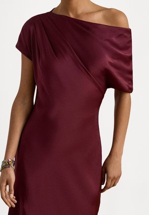 Femme portant une robe bordeaux en satin à une épaule avec une manche courte et un bracelet coloré en perles au poignet gauche.