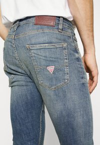 Vaqueros de mezclilla azul desgastados con un parche de cuero en la cintura, que presentan un logo triangular rojo y blanco en el bolsillo trasero.