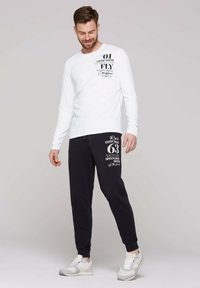 Camicia bianca a maniche lunghe con testo grafico nero abbinata a pantaloni sportivi neri con stampa bianca. Il modello indossa sneaker di colore chiaro.