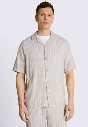 Mann trägt beige-weiß gestreiftes Kurzarmhemd mit passenden Hosen vor neutralem Hintergrund.