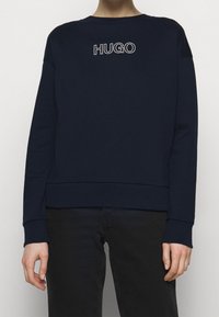 Mörkblå sweatshirt med rund halsringning, ribbade muddar och nederkant. Har en vit "HUGO"-logotyp på övre ryggen. Mjukt material och avslappnad passform.