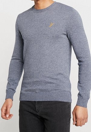 Mężczyzna ubrany w dopasowany jasnoszary sweter dziany z długimi rękawami i małym złotym haftowanym logo na piersi, w połączeniu z czarnymi dżinsami.