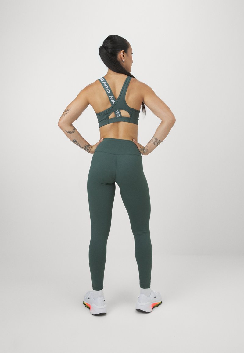 Reggiseno sportivo verde scuro con un design con spacchi e logo sulle cinghie, abbinato a leggings a vita alta coordinati e scarpe da ginnastica bianche.