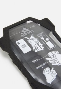 Porta telefono in tessuto nero con parte anteriore trasparente. Presenta istruzioni per l'uso e il logo Adidas. Progettato per smartphone e piccoli oggetti.