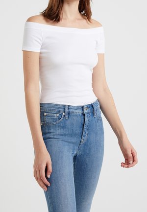 Femme portant un haut blanc ajusté sans épaules et un jean bleu taille haute sur un fond clair uni.