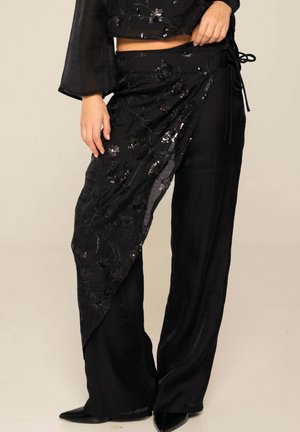 Pantalones negros de pierna ancha con detalle de envoltura con flores de lentejuelas en una pierna, combinados con top corto a juego y zapatos negros puntiagudos.