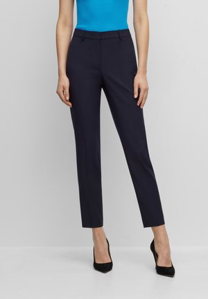 Femme debout portant un pantalon bleu marine ajusté longueur cheville, des talons hauts noirs pointus et un haut sans manches bleu vif.