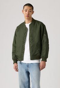 Ung mann med kort hår iført olivengrønn bomberjakke over hvit skjorte og lyseblå jeans, står foran en ensfarget bakgrunn.