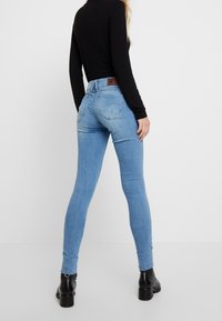 Jeans ajustados de mezclilla azul que presentan una cintura de altura media, bolsillos traseros y una etiqueta de cuero en la cinturilla. El modelo lleva botas negras hasta el tobillo.