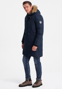 Námořnická parka s umělou kožešinou na kapuci, dvěma předními kapsami a zipem. Nositá přes džíny a hnědé turistické boty.