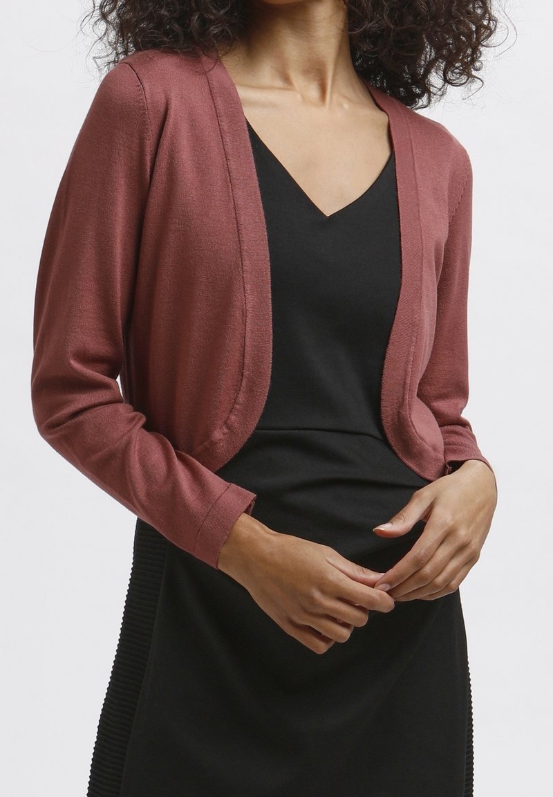 Femme portant un cardigan ouvert de couleur rouille sur une robe noire à col en V, les mains croisées devant elle.
