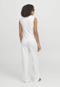 Biała bezrękawnik ze swobodnymi, szerokimi białymi spodniami. Gładki materiał z minimalnymi detalami szwów. Modelka stoi z kręconymi włosami.