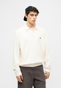 Νεαρός άντρας με λευκό μπλουζάκι πόλο μακρυμάνικο, σκούρο παντελόνι και μαύρο καπέλο γυρισμένο ανάποδα, στέκεται μπροστά σε απλό ανοιχτό φόντο.