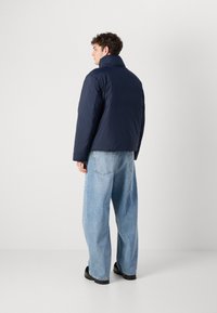 Marineblauwe pufferjack met een hoge kraag, gecombineerd met lichtblauwe oversized spijkerbroeken en zwarte laarzen. Soepele textuur, casual pasvorm.