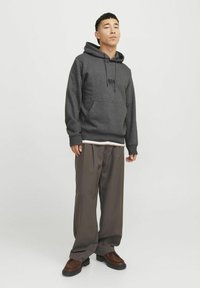 Sudadera gris con bolsillo frontal y logotipo negro, combinada con pantalones sueltos marrones y zapatos marrones. Diseño simple con un ajuste relajado.