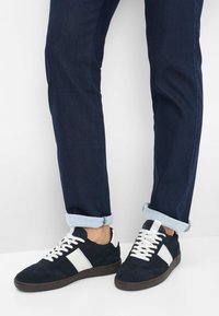 Donkerblauwe jeans met opgerolde pijpen, gecombineerd met zwarte sneakers met witte zijkanten en witte veters op een witte vloer.