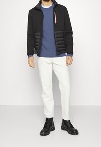 Black jacket with a padded front, blue textured sweater, white pants, and black lace-up boots. Jacket includes zip pocket with tricolor accent.