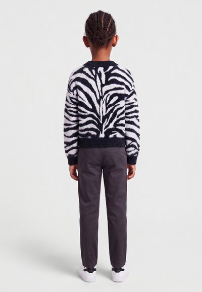 Enfant aux cheveux tressés portant un pull rayé zèbre noir et blanc, pantalon foncé et baskets blanches, debout de dos sur un fond uni.