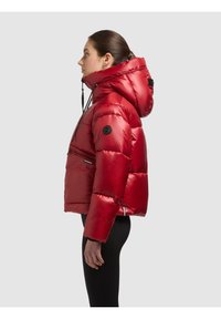 Profiilissa oleva nainen, joka pukeutunut kiiltävään punaiseen toppatakkiin, jossa on suuri huppu, ja mustiin housuihin, yksinkertaisen vaaleanharmaan taustan edessä.