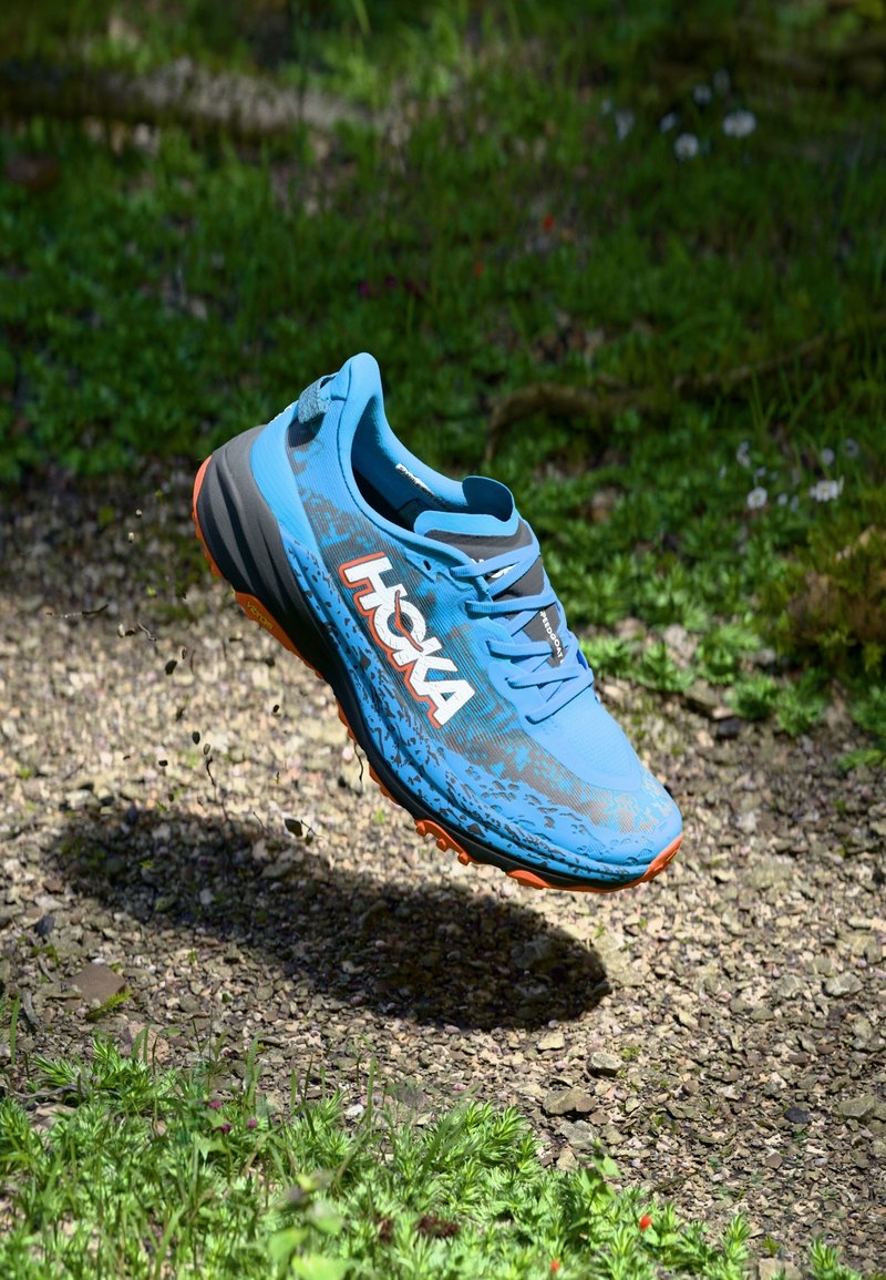 Chaussure de course bleue Hoka suspendue en l'air au-dessus du gravier et de l'herbe en extérieur, montrant le marquage latéral et des accents orange sur la semelle.