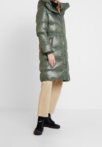 Grüner, glänzender Steppmantel mit Kapuze, ausgestattet mit einem Reißverschluss und gestepptem Muster. Kombiniert mit beigen Hosen und schwarzen Schnürstiefeln.