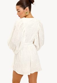 Witte jurk met pofmouwen, geborduurd bloemenpatroon, ingezette taille en textiel met textuur. Gouden oorbellen in de vorm van de zon complementeren het ontwerp.