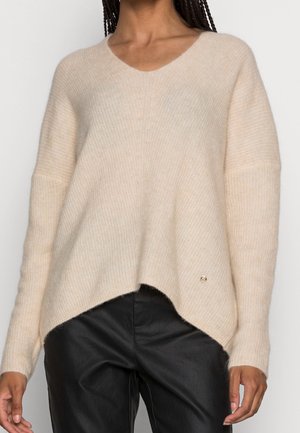 Woman wearing a loose, cream-colored knit sweater with a V-neck and asymmetrical hem paired with black leather pants.