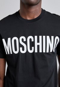 T-shirt in cotone nero con il logo "MOSCHINO" in bianco in evidenza sulla parte frontale. Design classico con collo rotondo e maniche corte.