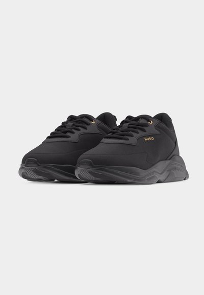 Paire de baskets noires Hugo avec semelles épaisses, lacets sur le devant, et petit logo doré HUGO sur le côté.
