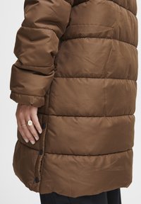 Brun oppustet frakke med quiltet design, der har høj krave, lange ærmer og sidelukninger med knapper ved kanten. Glat tekstur.