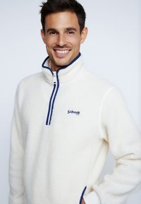 Fleece pullover i off-white med høj krave, halvlås og marineblå detaljer. Har logo på brystet og en blød, tekstureret overflade.
