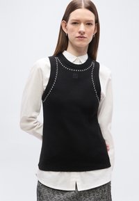 Νεαρή γυναίκα με ίσια καστανά μαλλιά φοράει μαύρο αμάνικο τοπ με ασημένιες τρουκς πάνω από λευκό πουκάμισο με κουμπιά και φούστα με σχέδιο.