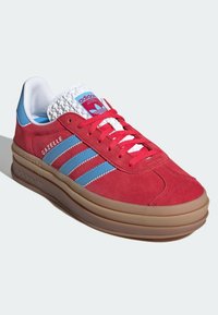 Červené semišové tenisky s modrými akcenty, bílým texturovaným jazykem a třemi horizontálními pruhy. Mají polstrovanou gumovou podrážku a značkovou detaily.