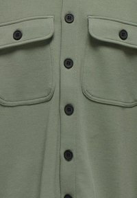Camicia verde strutturata con texture, caratterizzata da sei bottoni neri sulla parte anteriore e due tasche sul petto con patte.