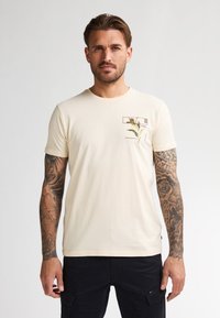 Camiseta de algodón color crema con cuello redondo, que presenta un pequeño gráfico de una planta en la parte izquierda del pecho. Mangas cortas y un corte relajado.