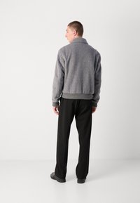 Gray herringbone jacket with a collar, ribbed cuffs, and waistband. Worn with black trousers and simple black shoes. Back view.