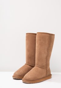 Par de botas de invierno de ante marrón hasta la mitad de la pantorrilla, con suelas de goma planas, costuras visibles en los lados y forro de felpa en el interior.