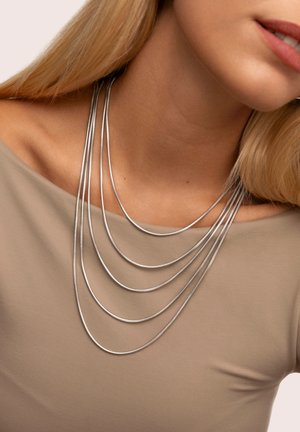 Sömlös, flerlagrig silverhalsband med en slät och mjuk textur, buren över en mjuk beige topp med båtformad neckline.